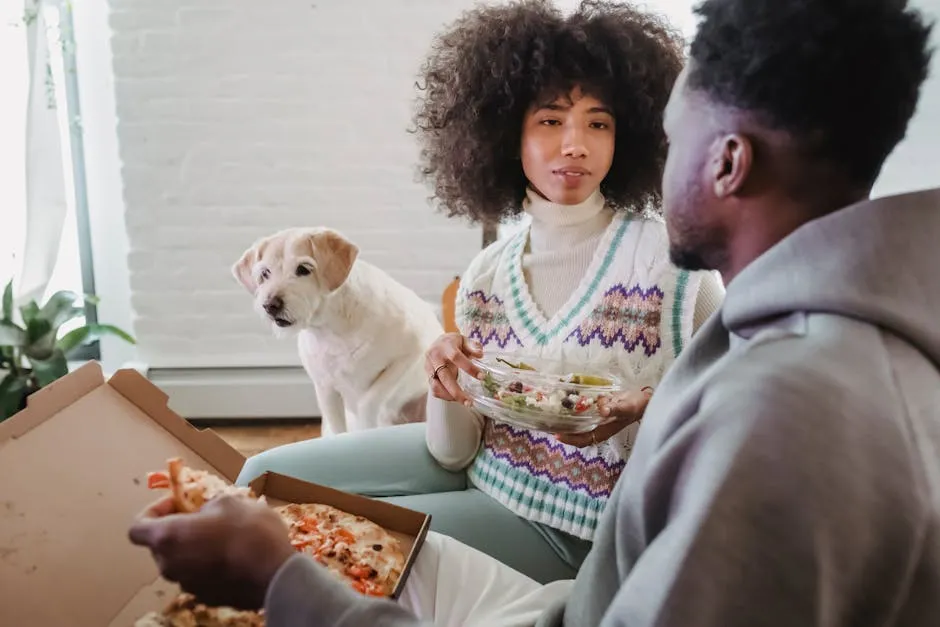 Crop African American couple speaking with each other while having snack with pizza and salad at home with hungry dog