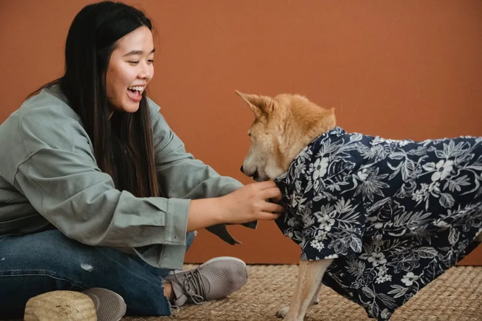 Side view of expressive Asian female owner dressing dog while sitting with crossed legs on floor
