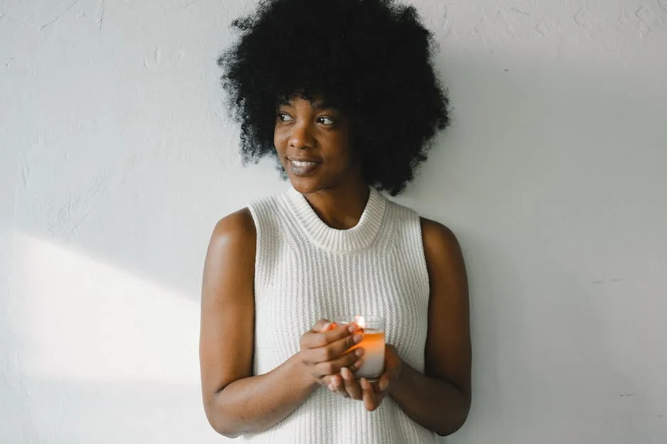 Gentle African American female with curly hair standing with aromatic burning candle in glass at home and looking away