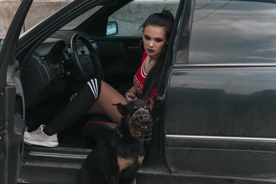 A stylish young woman sits in a car with a muzzled dog at her side, showcasing urban fashion.