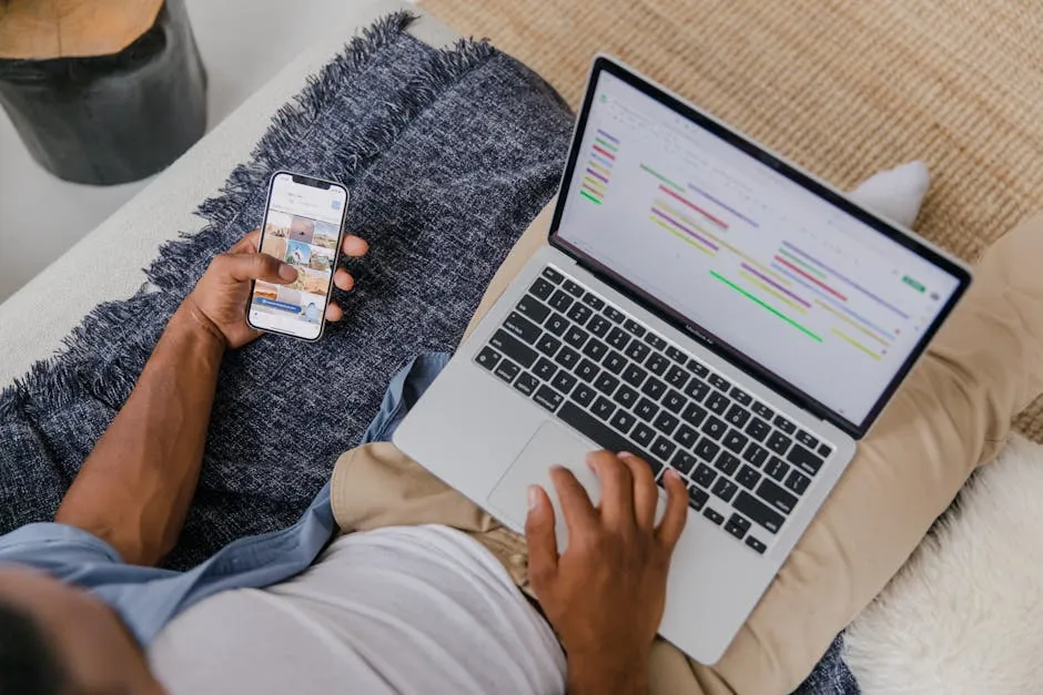 Man multitasking with phone and laptop on sofa, showcasing modern remote work lifestyle.